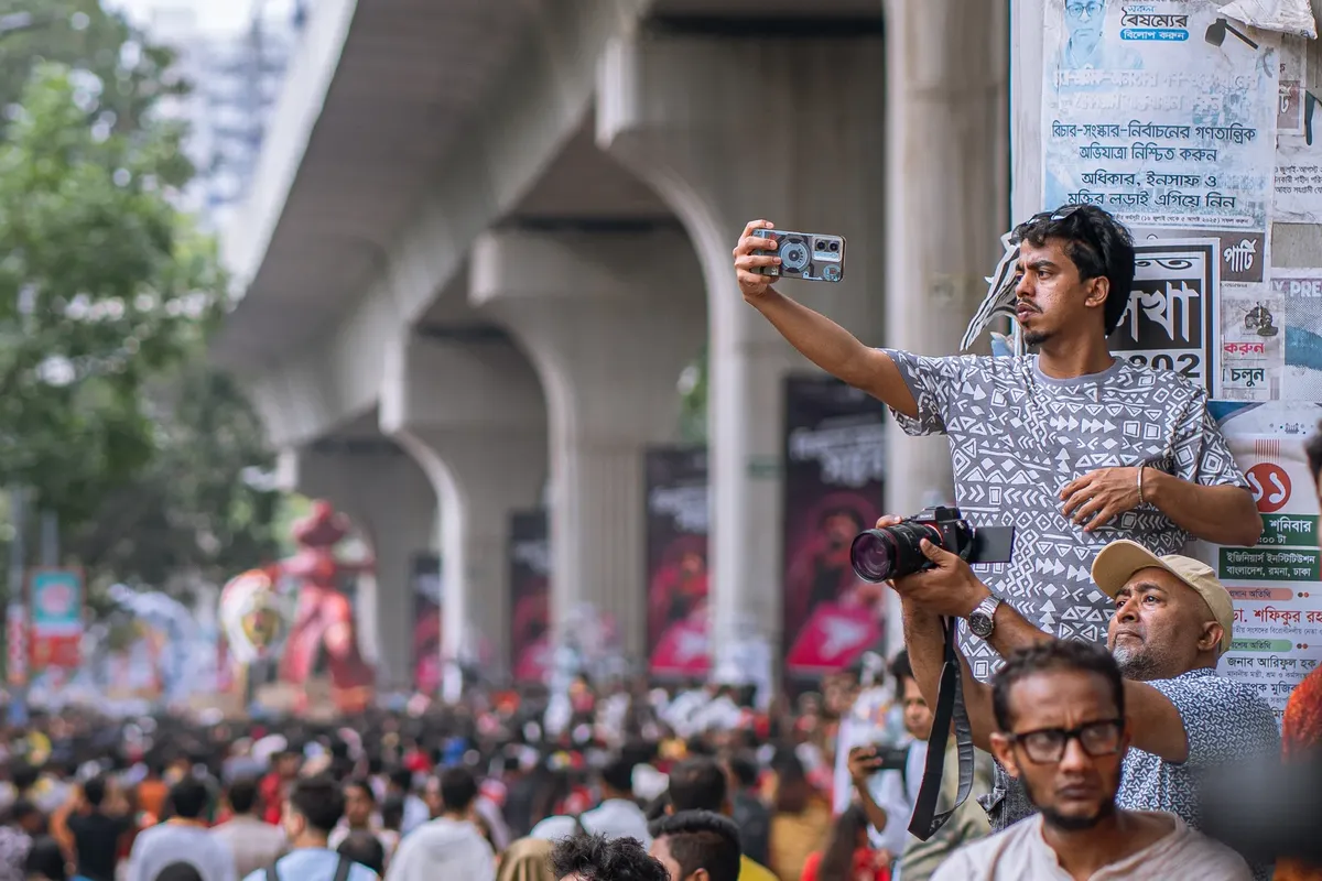A lively street festival with a crowd capturing moments on camera amid colorful decorations and urban infrastructure.