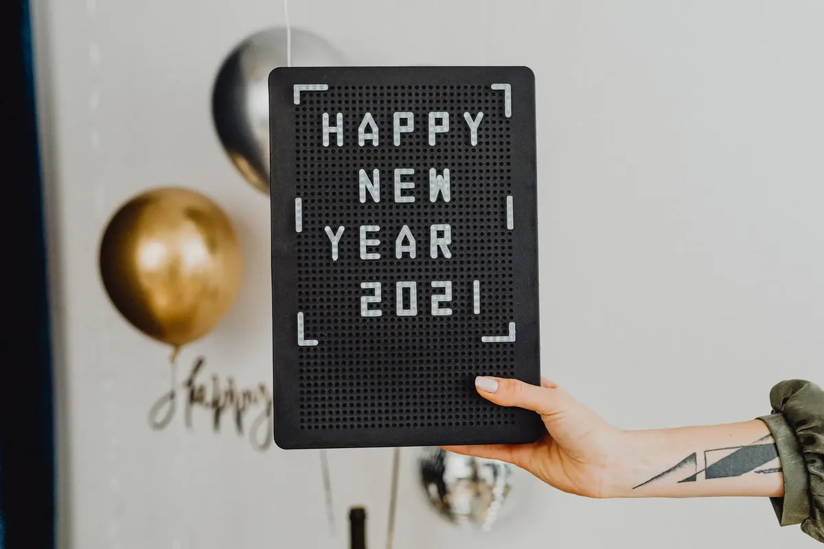 Hand holding a letter board with 'Happy New Year 2021' message, adorned by balloons in the background.