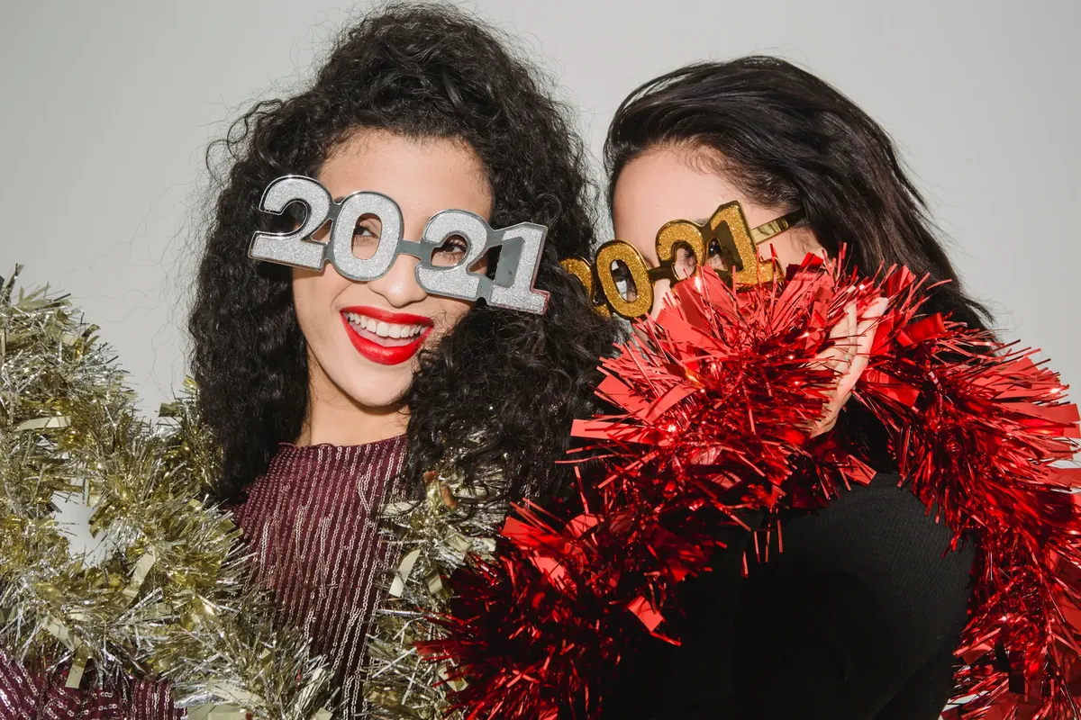 Women celebrating New Year's Eve with 2021 glasses and tinsel indoors, showcasing joyful expressions.